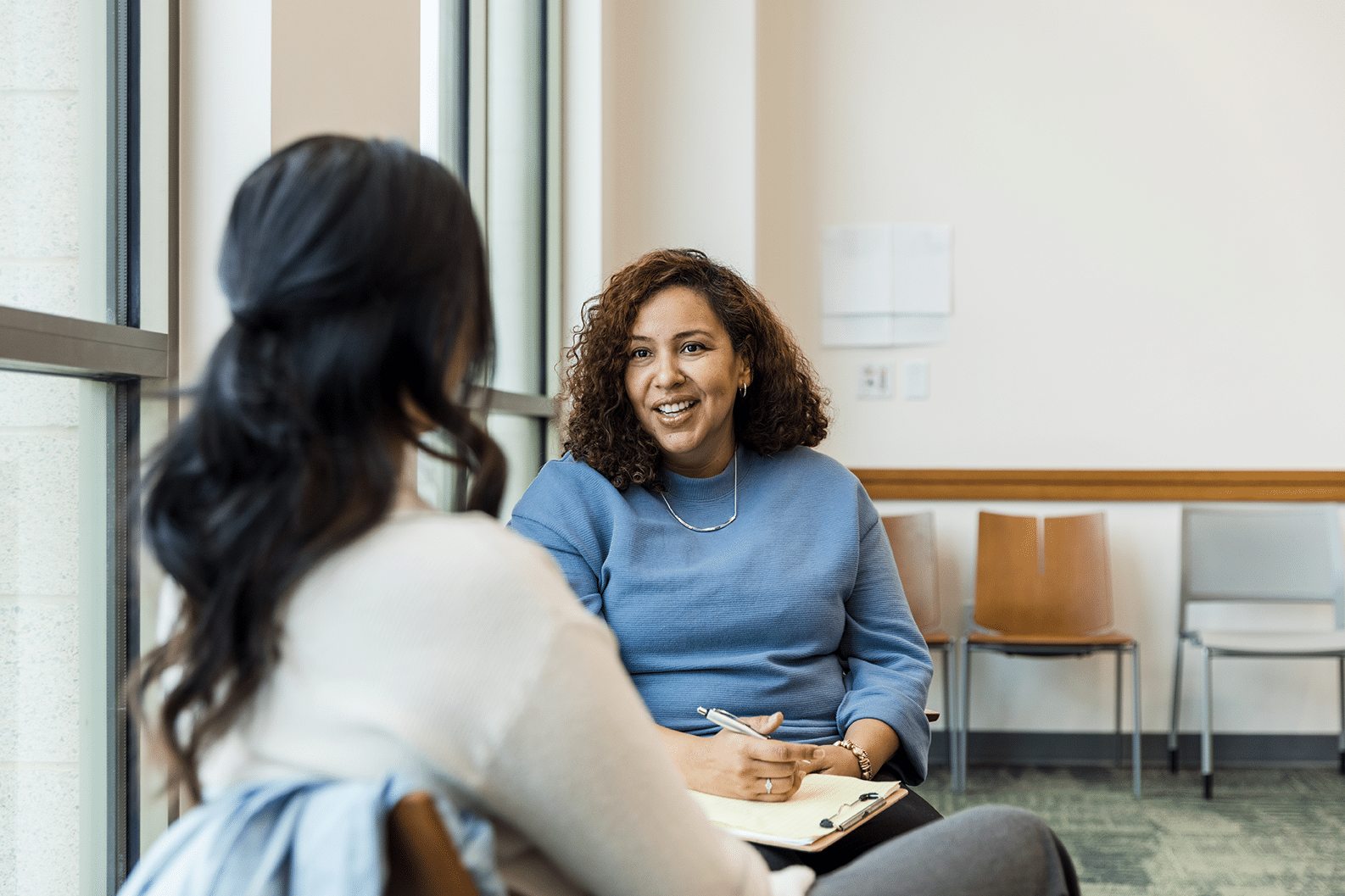 Seorang wanita yang memegang buku catatan tersenyum sambil berbincang dengan seorang wanita yang duduk di hadapannya di sebuah kantor yang terang benderang.